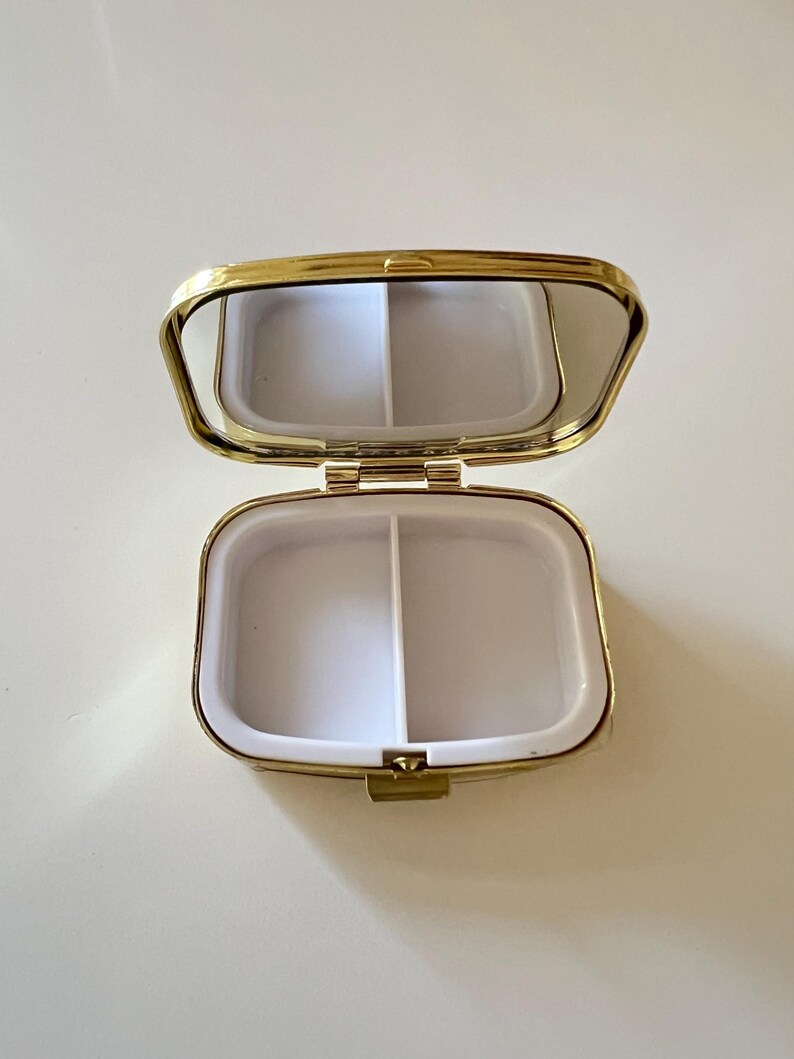 May include: Open, gold-toned pill box with a mirrored lid. The interior is divided into two white compartments. The pill box is rectangular with rounded edges and a clasp closure.