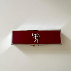 May include: A rectangular metal case with a glossy deep red enamel finish and a silver-toned border. Centered on the front is a small, raised silver-toned emblem depicting a winged cherub figure holding a bow and arrow in a profile stance.
