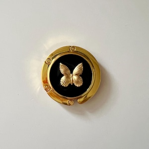 May include: A circular gold-tone metal bag hanger featuring a black center inlay. A three-dimensional gold butterfly is centered on the black surface. The hanger has a hinged design with visible metal joints along the outer circular frame.