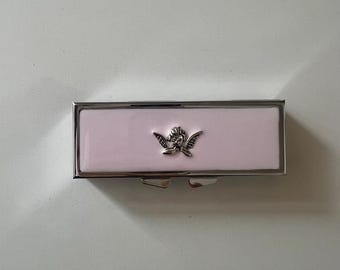 Könnte beinhalten: Ein rechteckiges Metallgehäuse mit einer glatten, hellrosa Emaille-Oberfläche und einem silberfarbenen Rahmen. In der Mitte der Vorderseite befindet sich ein kleines, detailliertes silberfarbenes Metallemblem eines geflügelten Putto.
