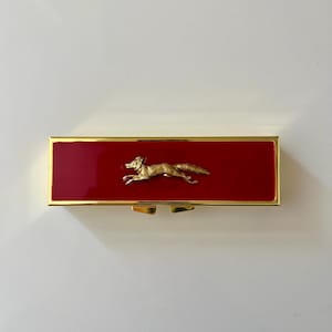 May include: A rectangular lipstick case featuring a glossy red enamel finish on the front panel, framed by a polished gold-tone metal border. A small, three-dimensional gold-tone metal figure of a running fox is centered on the red surface.