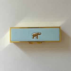 May include: A rectangular metal case with a light blue enamel finish and a gold-toned frame. The center of the lid features a small, embossed gold-colored elephant figure. The case has a hinged closure mechanism visible along the bottom edge.