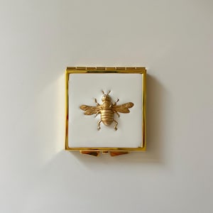 Espejo de bolsillo con forma de abeja: Espejo compacto de metal dorado con incrustaciones de resina blanca y abeja de latón.