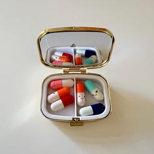 May include: A small, open pill box with a gold-colored frame and a mirrored lid. The box is divided into two compartments, each containing colorful, cartoon-style pills in shades of red, orange, pink, blue, and green. Some pills have smiling faces.