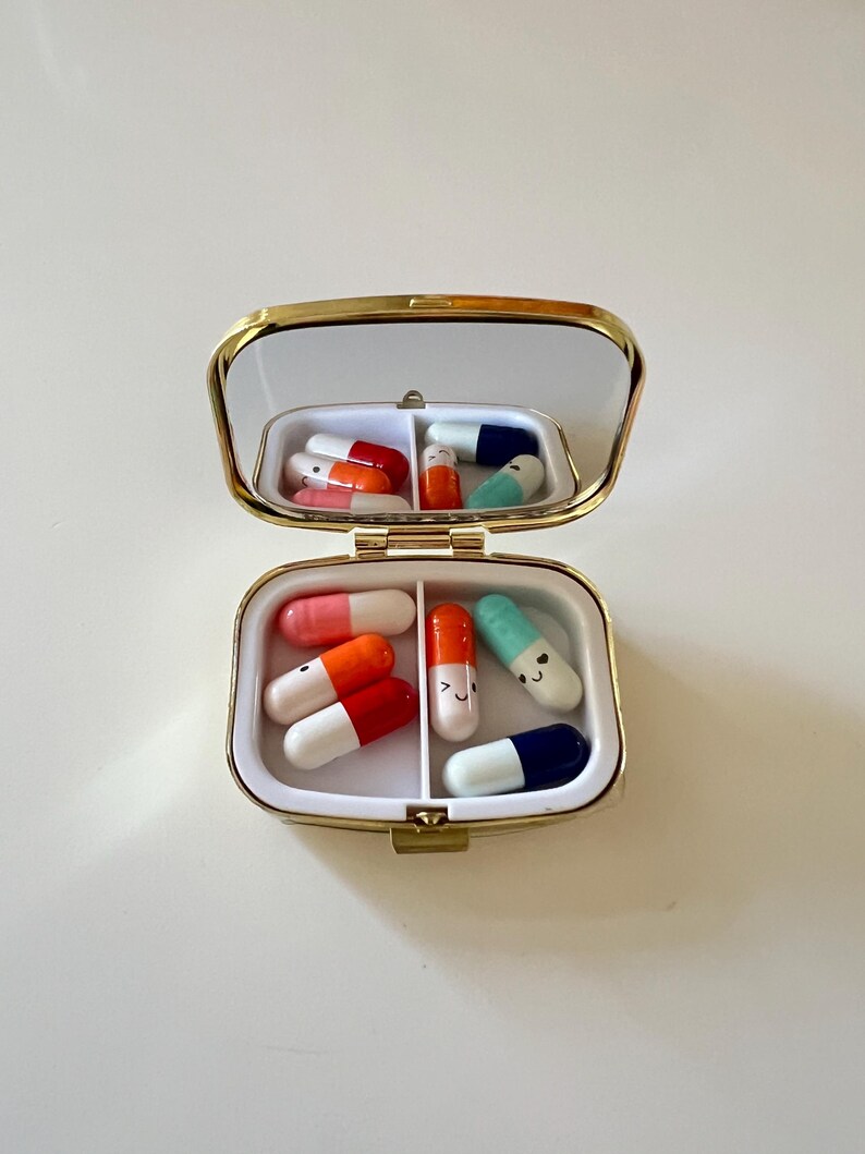 May include: A small, open pill box with a gold-colored frame and a mirrored lid. The box is divided into two compartments, each containing colorful, cartoon-style pills in shades of red, orange, pink, blue, and green. Some pills have smiling faces.