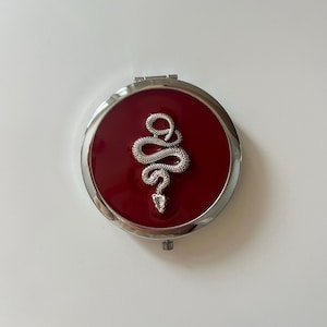 May include: A round, silver-toned compact mirror featuring a deep red, glossy enamel inlay on the lid. Centered on the red surface is a textured, silver-colored snake emblem with a coiled body and a detailed head facing downward.