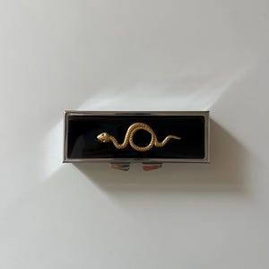 May include: A small, rectangular silver compact with a black enamel surface. A gold-colored snake design is centered on the compact. The compact is closed and sits on a white surface.