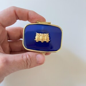 May include: A rectangular, hinged trinket box with a deep blue enamel finish and gold-tone trim. The lid features a gold-colored relief of three owls. The box is held in a hand against a white background.