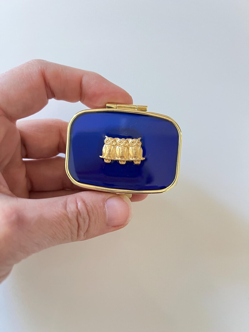 May include: A rectangular, hinged trinket box with a deep blue enamel finish and gold-tone trim. The lid features a gold-colored relief of three owls. The box is held in a hand against a white background.
