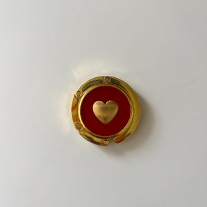 May include: A circular gold-tone metal bag hanger featuring a vibrant red circular inlay at the center. A small, polished gold-tone heart shape is positioned directly in the middle of the red inlay, creating a distinct contrast against the metallic frame.