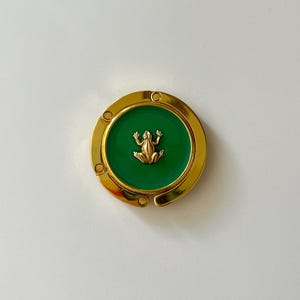 May include: A circular gold-tone metal bag hanger featuring a vibrant green enamel inlay at the center. A small, three-dimensional gold-colored frog figure is positioned in the middle of the green circle. The hanger has a hinged design with visible joints.