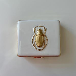 May include: A white, rectangular trinket box with a gold-colored scarab beetle design on the lid. The beetle is in the center of the box, and the box has gold trim. The box is on a white surface.