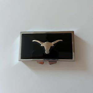 Op de afbeelding: Een zilveren en zwarte pillendoos met een longhorn-stierenkopontwerp.