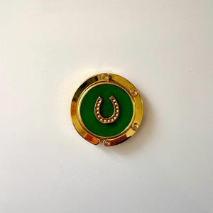 May include: A round, gold-toned metal bag hanger featuring a circular green enamel inlay. Centered on the green surface is a small, gold-toned horseshoe emblem with a textured, dotted finish. The outer gold frame has a smooth, polished appearance.