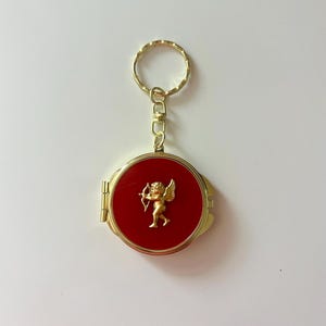 May include: A round, gold-toned metal compact mirror keychain featuring a vibrant red circular inlay on the front. A small, raised gold-colored figure of a winged cherub holding a bow and arrow is centered on the red surface. The item includes a gold metal ring.