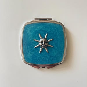 May include: A square compact mirror with a metallic silver frame and a teal, glittery resin inlay. Centered on the teal surface is a three-dimensional silver-toned sun emblem featuring a human face with rays extending outward from the center.