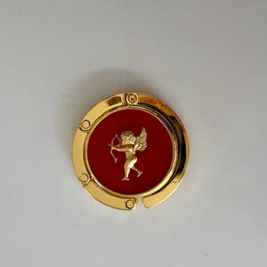 May include: A gold-colored, circular purse hook with a red center. The center features a gold-colored cupid with a bow and arrow. The hook has a hinged design and is decorated with small, round rivets.