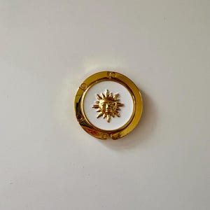 May include: A gold-colored, circular decorative element featuring a white center with a sun face design. The sun has a smiling face and radiating lines. The outer rim is gold with small decorative accents. The item is likely a furniture accent or decorative piece.