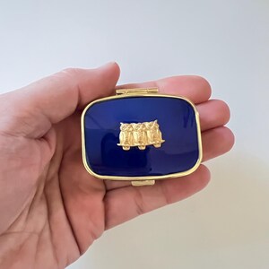 May include: A rectangular, navy blue pillbox with a gold-colored frame. The lid features a gold-colored relief of three owls. The pillbox is held in a hand against a white background.