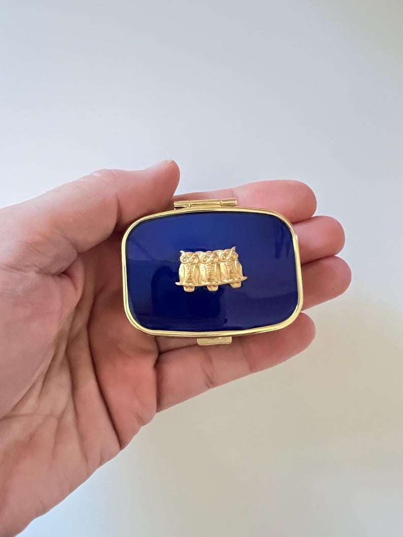 May include: A rectangular, navy blue pillbox with a gold-colored frame. The lid features a gold-colored relief of three owls. The pillbox is held in a hand against a white background.