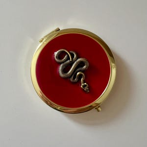 May include: A gold compact mirror with a red lid and a gold snake design.