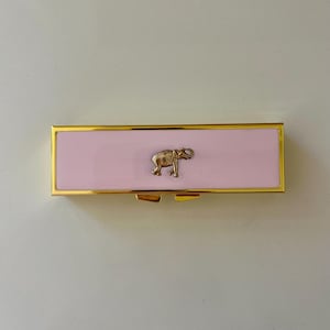 May include: A rectangular lipstick case featuring a smooth, light pink enamel finish framed by a polished gold-tone metal border. A small, detailed gold-tone metal elephant emblem is centered on the front surface of the case.