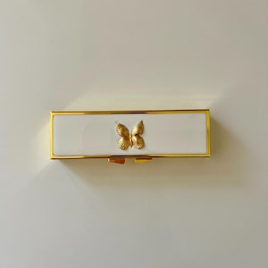 May include: A rectangular white box with gold trim and a gold butterfly emblem. The box has a hinged lid and a gold clasp. The butterfly is centered on the lid.