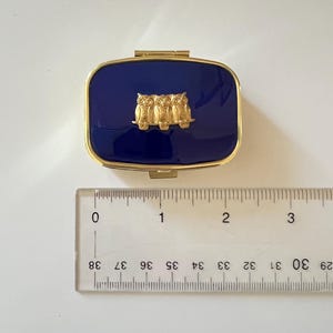 May include: A rectangular, navy blue pillbox with a gold-colored frame and clasp. The lid features a gold-colored relief of three owls. The pillbox measures approximately 1.5 inches long.