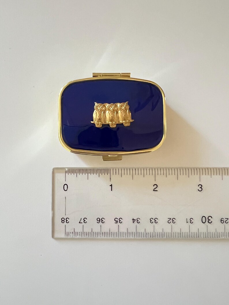 May include: A rectangular, navy blue pillbox with a gold-colored frame and clasp. The lid features a gold-colored relief of three owls. The pillbox measures approximately 1.5 inches long.