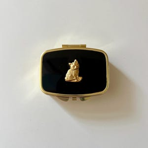 May include: A rectangular gold-tone metal pill box with a glossy black enamel lid. The center of the lid features a small, raised gold-tone cat figurine in a seated position. The box has a hinged closure and a polished metallic finish.