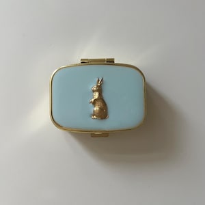 以下が含まれることがあります： 長方形の宝石箱。淡いブルーのエナメル蓋とゴールドのトリム。ゴールドのウサギの置物が蓋の中央に配置されています。箱は閉じられており、白い表面に置かれています。