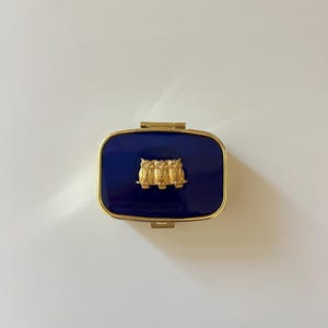 A small gold pill box with a push button latch. The top is coated with navy blue resin and has three tiny brass owls set in the center.