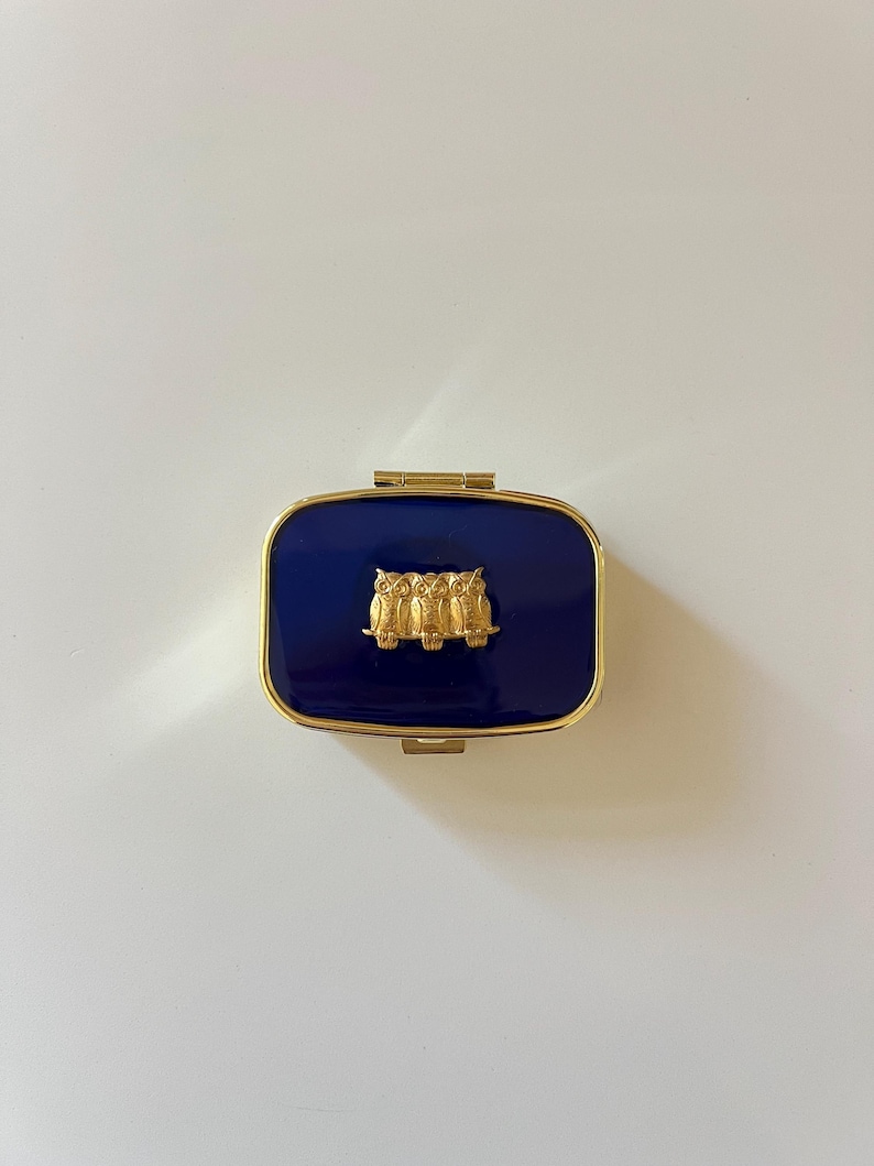 A small gold pill box with a push button latch. The top is coated with navy blue resin and has three tiny brass owls set in the center.