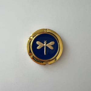 May include: A gold-colored purse hook with a navy blue center. The center features a gold dragonfly design. The hook is round with a hinged design for hanging purses and bags. The hook is on a white surface.
