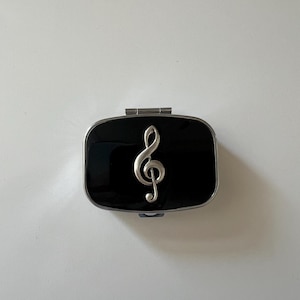 May include: A black pill box with a silver treble clef design. The rectangular box has a shiny black surface and a silver metal frame. The treble clef is in the center of the box.