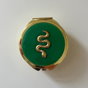 May include: A round gold-tone metal compact mirror featuring a vibrant green enamel inlay on the lid. Centered on the green surface is a raised, textured gold-tone snake emblem. The compact has a polished gold finish around the edges and a hinge.