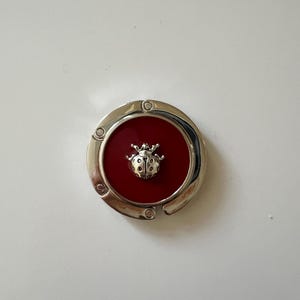 May include: A circular silver-toned metal bag hanger featuring a deep red center inlay. A small, three-dimensional silver-toned ladybug figure is centered on the red surface. The hanger has a hinged design with a smooth, polished metallic finish.