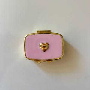 May include: A rectangular pill box with a glossy light pink enamel finish and a gold-toned metal frame. The center of the lid features a small, raised, polished gold-toned heart emblem. The container has a hinged lid and a secure latch closure.