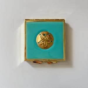 May include: A square compact mirror with a polished gold-tone metal frame and a vibrant turquoise enamel inlay. Centered on the turquoise surface is a raised, textured gold-tone sand dollar emblem featuring a classic five-petal star pattern.