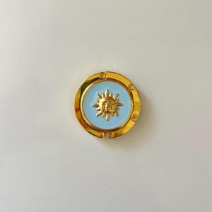 May include: A circular gold-tone metal bag hanger featuring a light blue enamel inlay. The center of the disc displays a raised gold sun face with radiating rays. The outer ring has a hinged design with small circular accents along the frame.