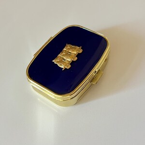 May include: A rectangular, gold-toned pill box with a navy blue enamel top. A gold-colored emblem is centered on the lid. The box has a hinged lid and a gold-colored frame.