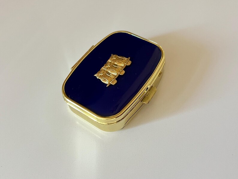 May include: A rectangular, gold-toned pill box with a navy blue enamel top. A gold-colored emblem is centered on the lid. The box has a hinged lid and a gold-colored frame.