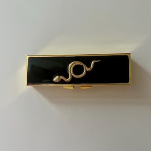May include: A rectangular lipstick case with a glossy black finish and a gold-toned metal trim. A textured gold-toned snake emblem is centered on the front surface of the case. The case features a hinged closure with visible gold-toned metal tabs.
