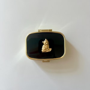 May include: A rectangular black compact with a gold-colored frame and clasp. A gold-colored fox figure is centered on the black surface. The compact is closed and sits on a white surface.