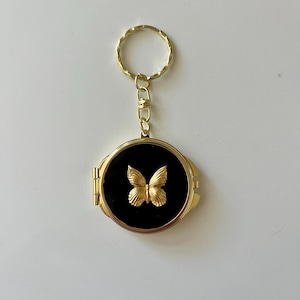 May include: A round gold-tone metal locket keychain featuring a black circular inlay on the front. A three-dimensional gold-tone butterfly emblem is centered on the black surface. The locket is attached to a gold-tone metal split ring by a short chain.