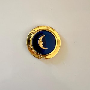 May include: A circular gold-tone metal bag hanger featuring a deep blue enamel center inlay. The center of the blue inlay displays a raised gold-tone crescent moon with a human face profile. The outer gold frame has a smooth, polished finish.