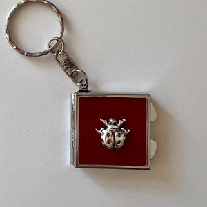 May include: A silver-colored keychain with a square, hinged compartment. The compartment has a red interior and a silver ladybug charm. The keychain includes a split ring and a lobster clasp.