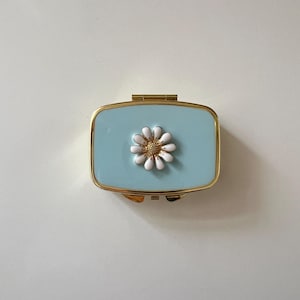 May include: A rectangular, light blue pill box with a gold-colored frame. A white and gold daisy is centered on the lid. The box is closed and sits on a white surface.