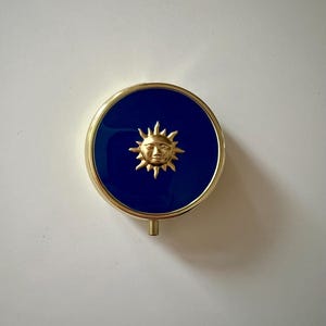 May include: A round, gold-toned compact mirror with a deep blue enamel center. The center features a gold sunburst design with a smiling face. The compact has a small, rectangular tab at the bottom.