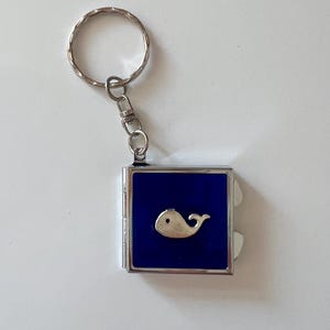 May include: A silver whale keychain with a blue background. The whale is a simple, stylized design. The keychain has a silver ring and clasp.